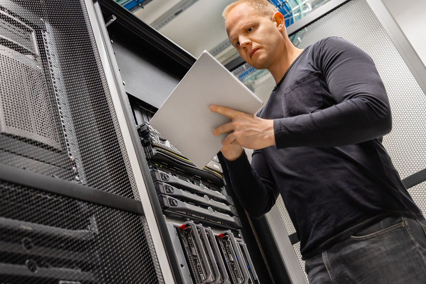 Confident male IT engineer using digital tablet standing in datacenter and monitoring servers and network.